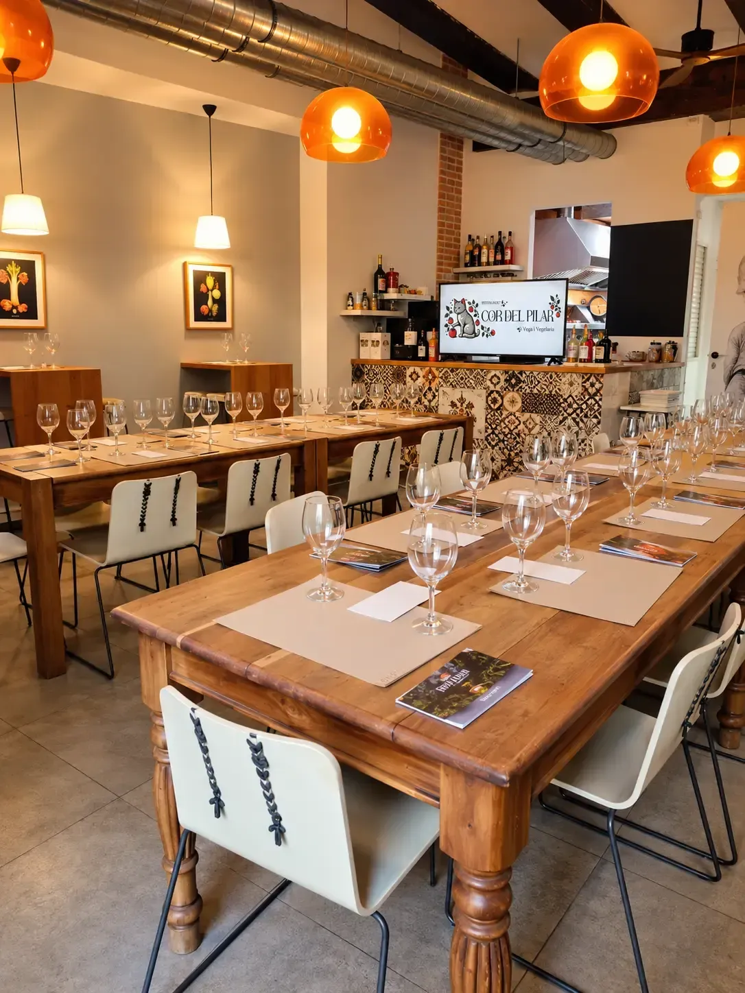 Acogedor comedor de restaurante con mesas puestas, luces colgantes de color naranja y una larga mesa comunal de madera.