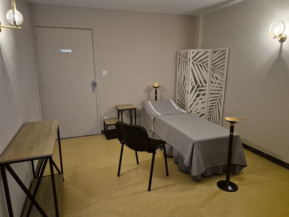 Salle de massage avec lit, paravent et éclairage d'ambiance ; tons beiges et gris.