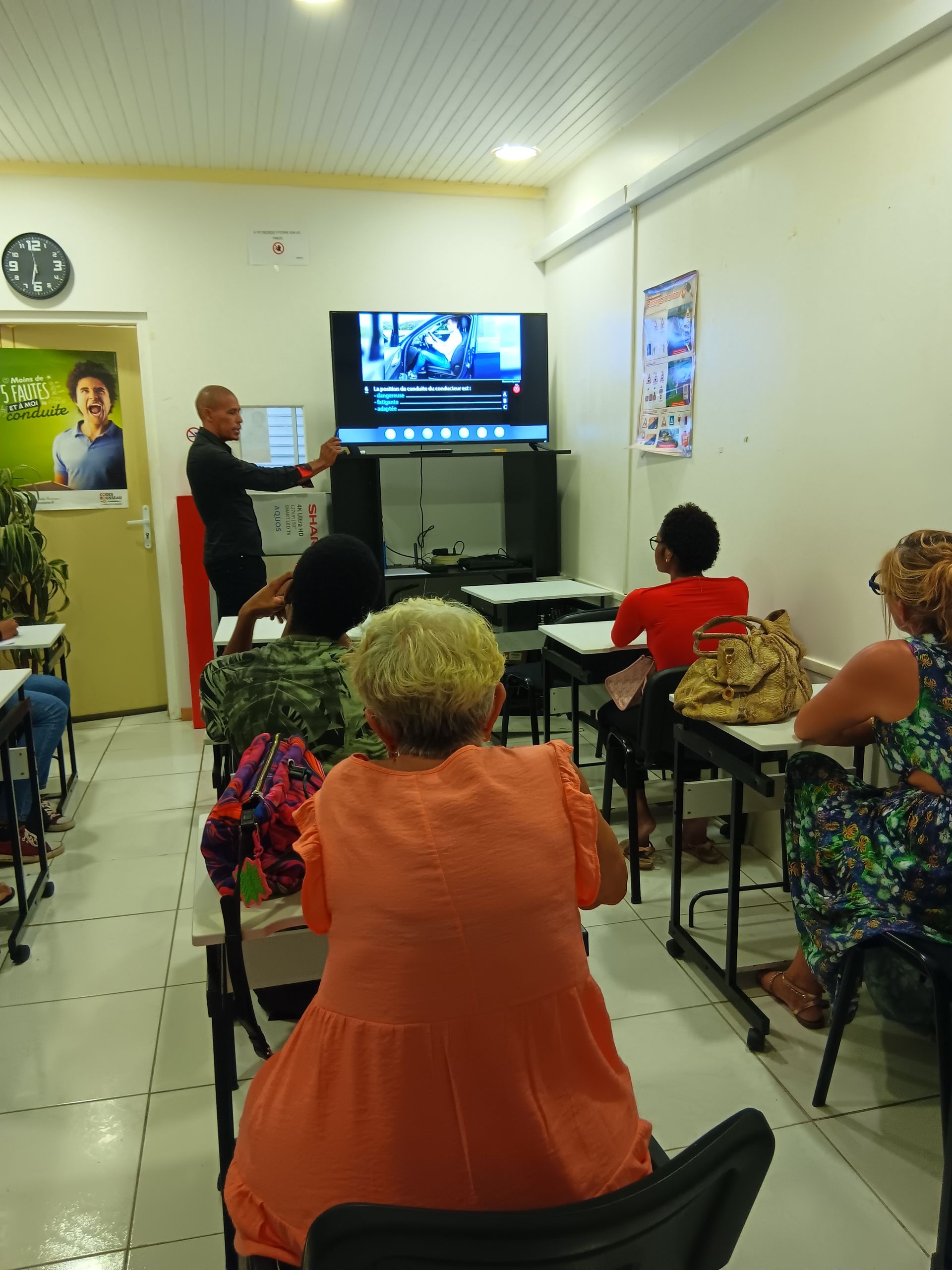 Un homme fait une présentation devant une petite classe, en désignant un écran diffusant une vidéo. Les élèves sont assis à leurs pupitres et regardent.