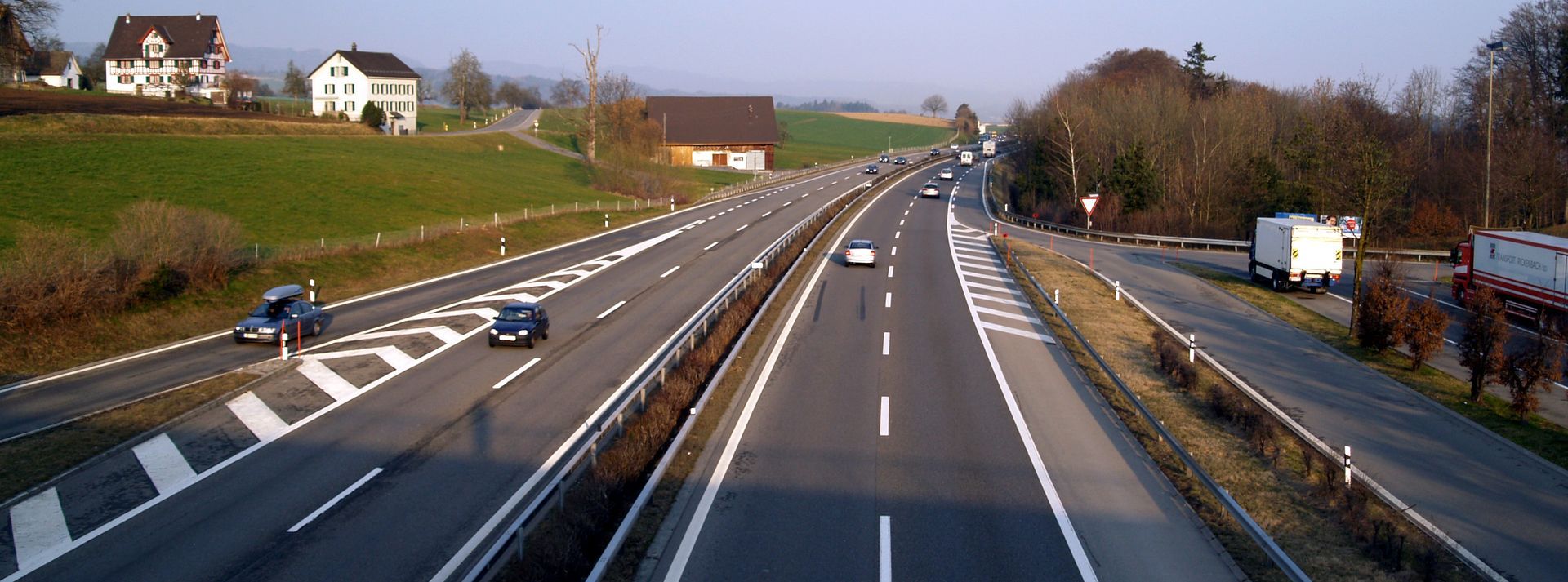 Autoroute à plusieurs voies, longeant des champs verdoyants et des bâtiments.