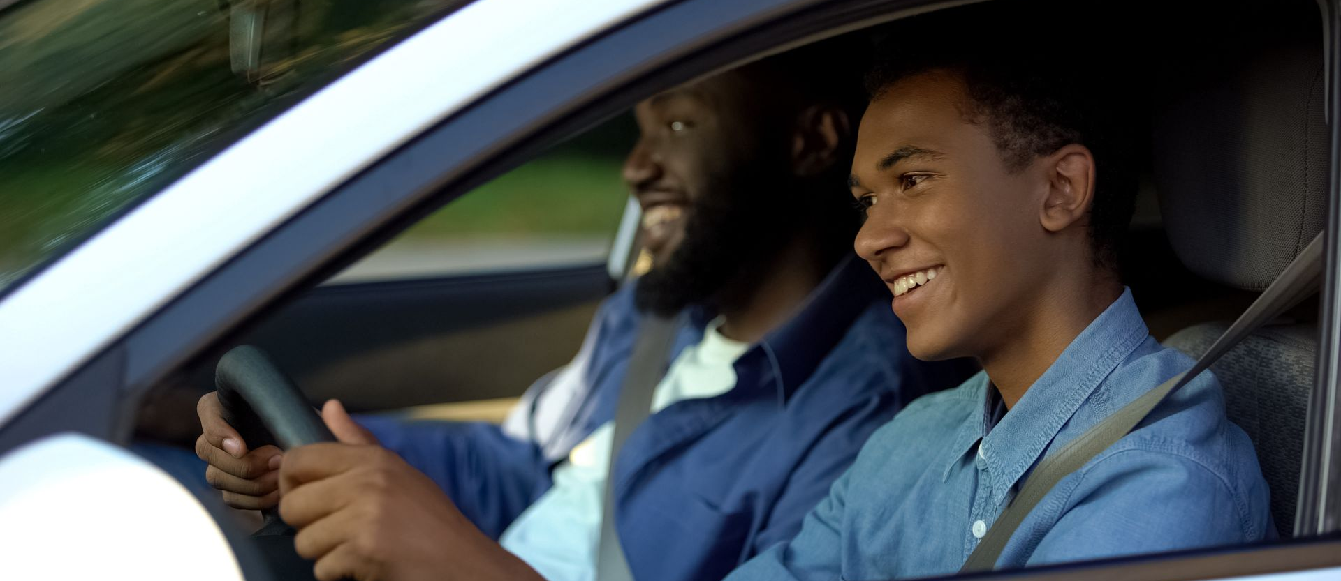 Deux personnes sourient dans une voiture. L'une d'elles tient le volant. Toutes deux portent des chemises bleues.