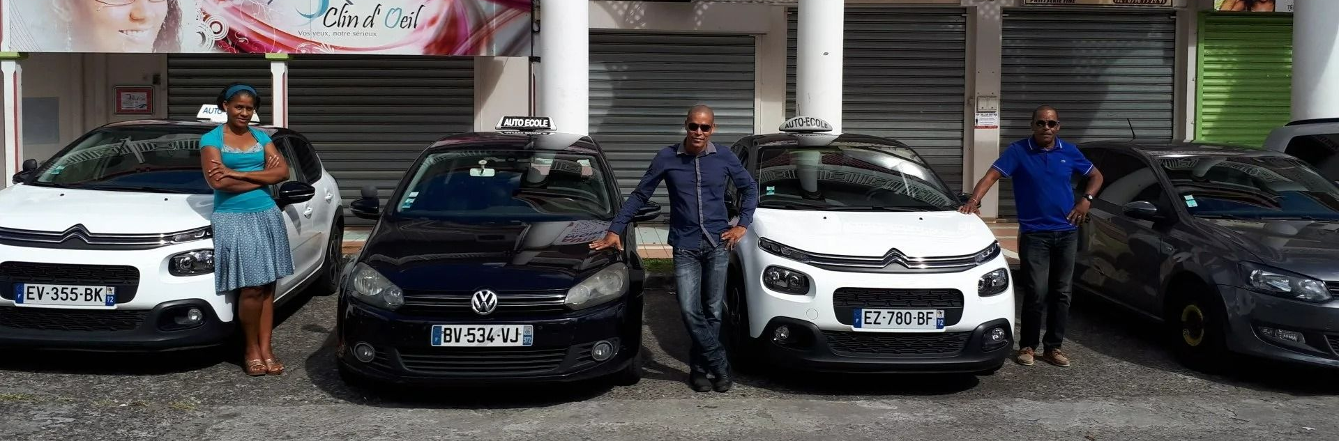 Quatre voitures avec des personnes posant pour la photo. Les voitures sont blanches, noires et gris foncé. Elles sont garées devant des vitrines de magasins.