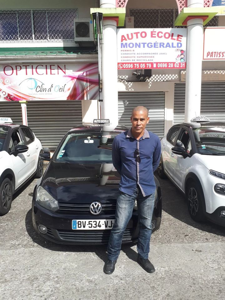 Un homme se tient devant une voiture noire, deux voitures blanches et un panneau d'auto-école.