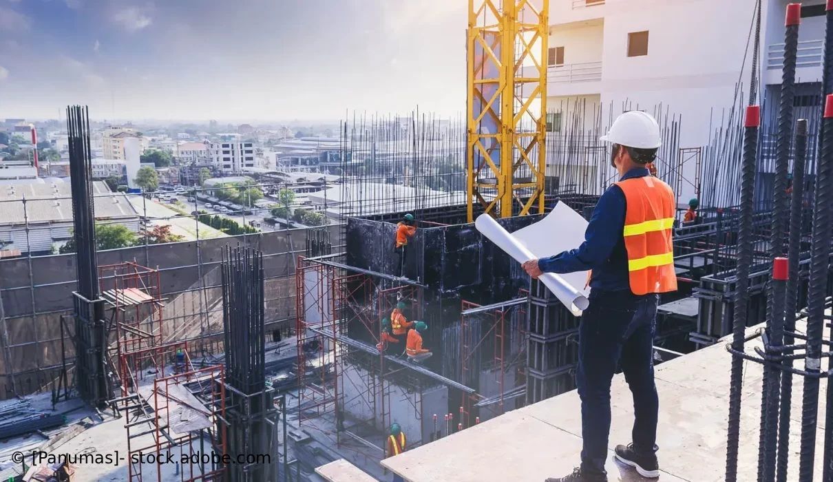 Ein Bauarbeiter mit Schutzhelm und Warnweste überwacht von einem Gerüst aus eine Hochhausbaustelle.