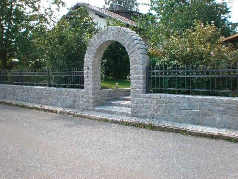 Ein steinernes Tor mit Bogen in einer niedrigen Steinmauer neben einer ruhigen Straße, dahinter Bäume und ein Haus