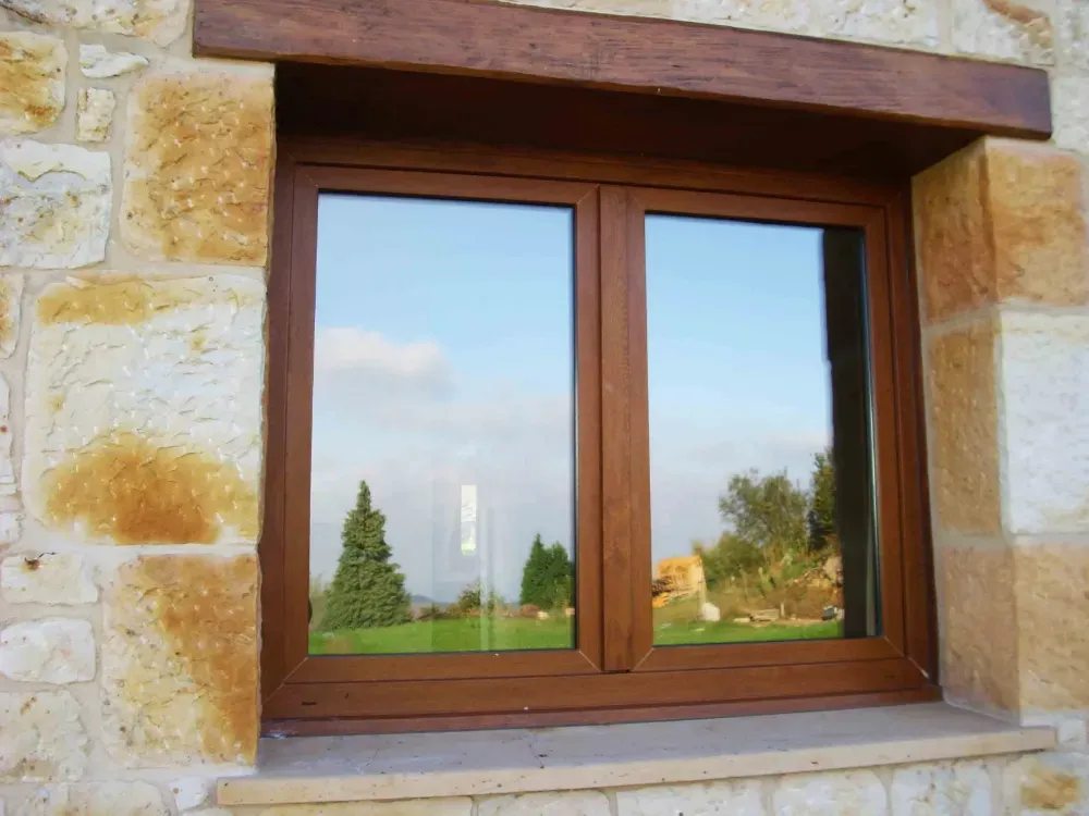 Ventana de madera empotrada en un muro de piedra, que refleja un paisaje verde y un cielo azul