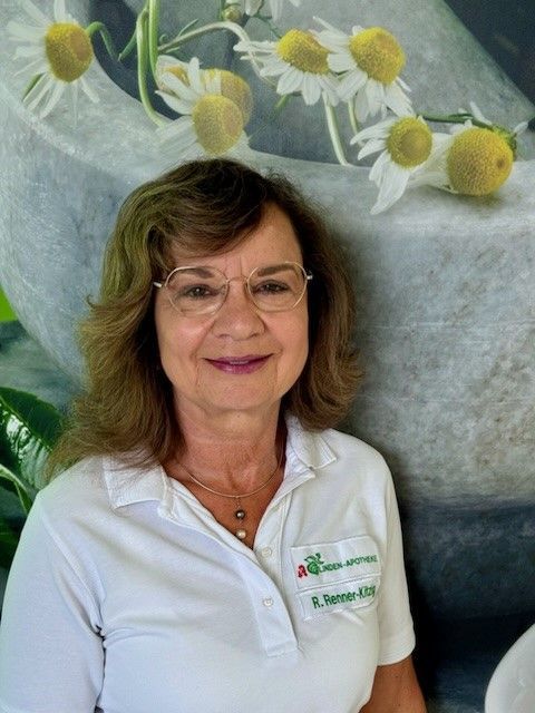 Eine Frau mit Brille und weißem Hemd lächelt vor einem Bild mit Gänseblümchen.