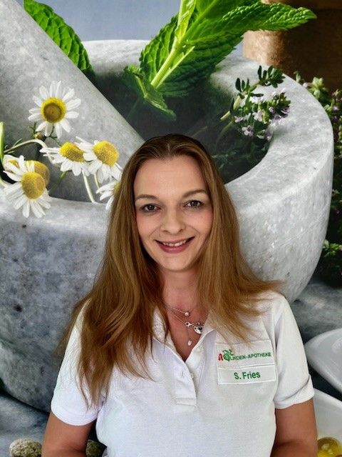 Eine Frau in einem weißen Hemd lächelt vor einem Bild mit Blumen und Kräutern.