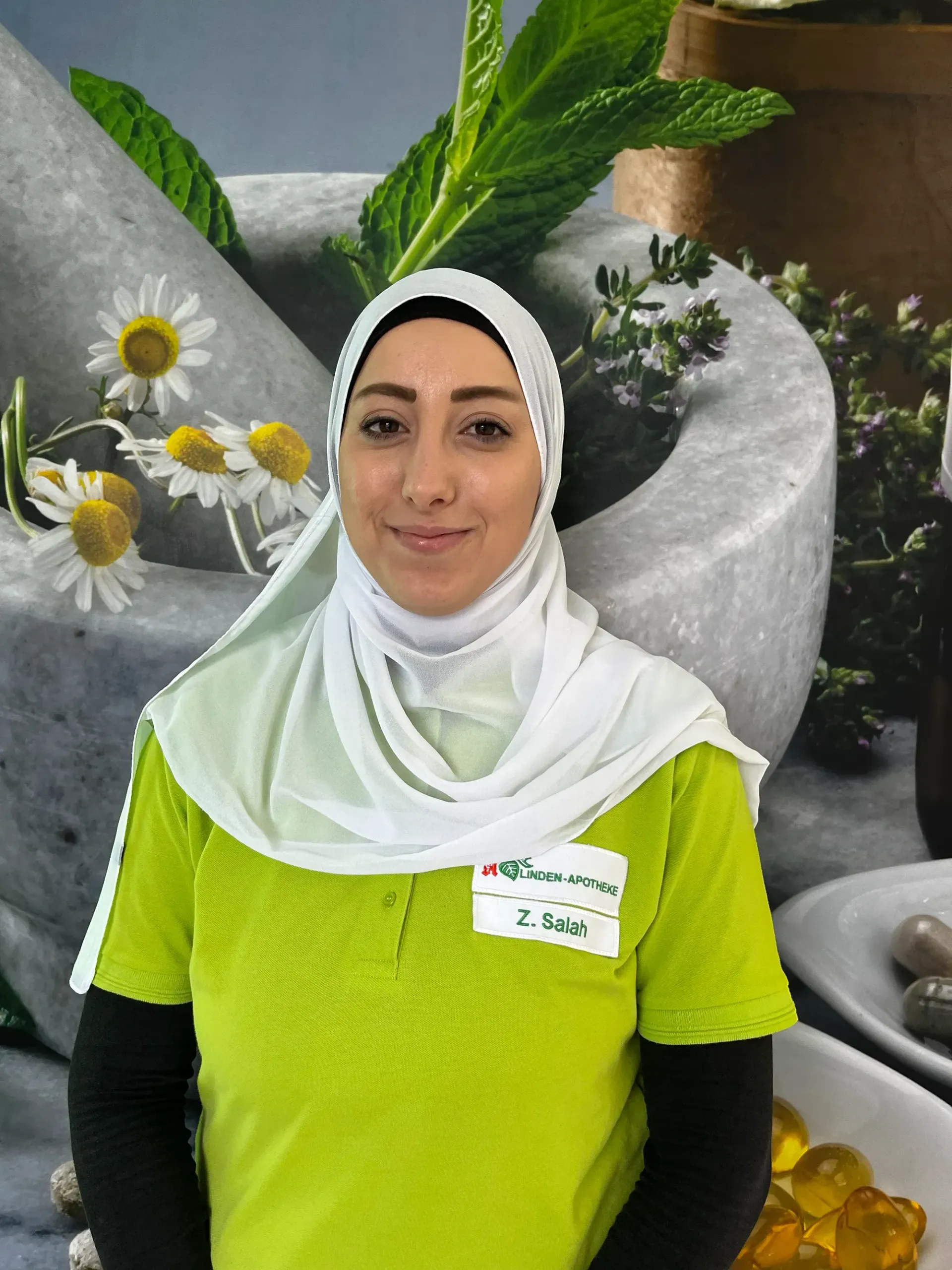 Frau in grünem Hemd und weißem Kopftuch lächelt, steht vor Mörser und Stößel mit Pflanzen.