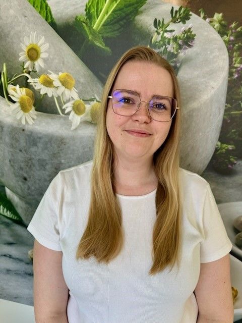 Eine Frau mit Brille und weißem Hemd steht vor einem Mörser und Stößel. Im Hintergrund sind Blumen zu sehen.