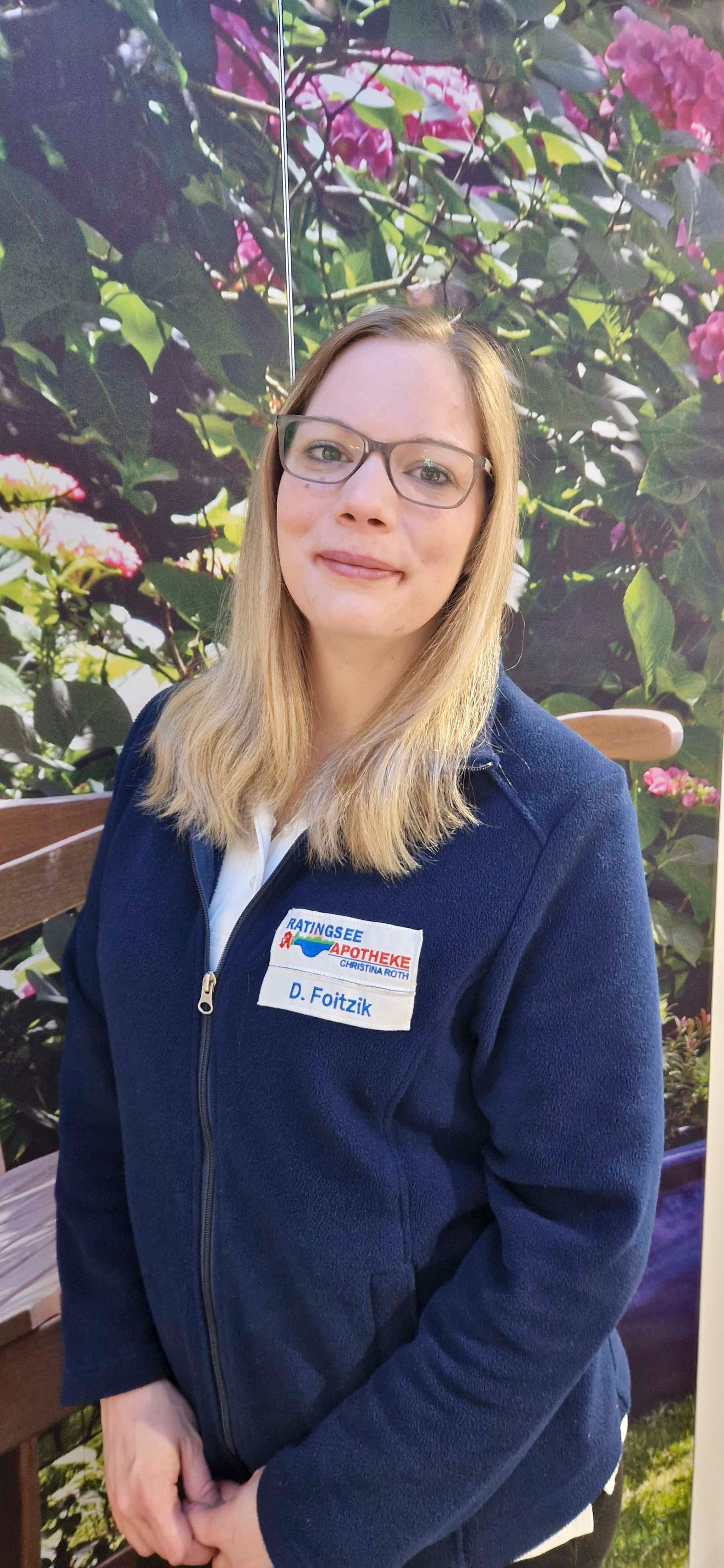 Eine Frau mit Brille und blauer Jacke steht vor einer Blumenwand.