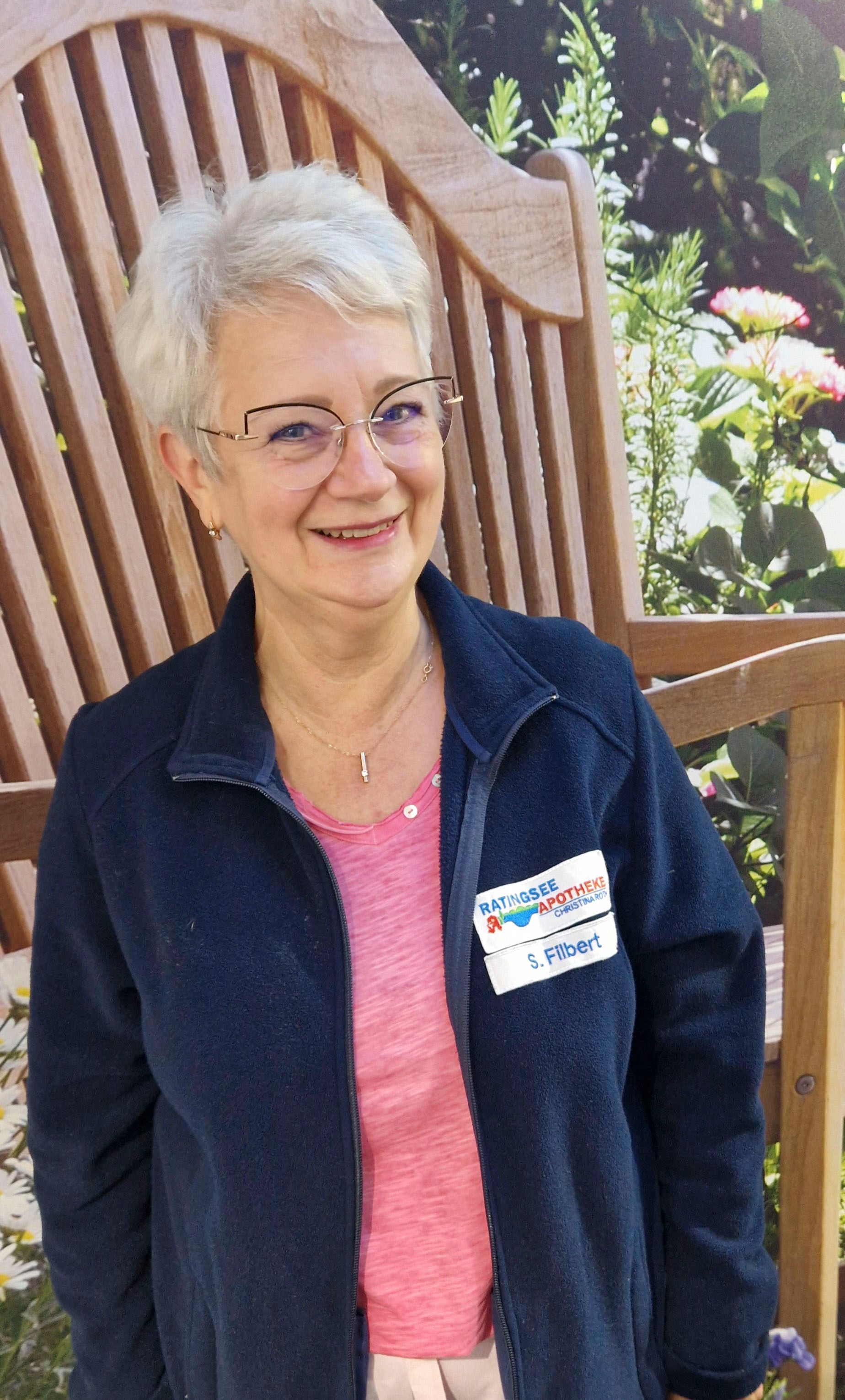 Eine Frau mit Brille und blauer Jacke steht vor einer Holzbank.