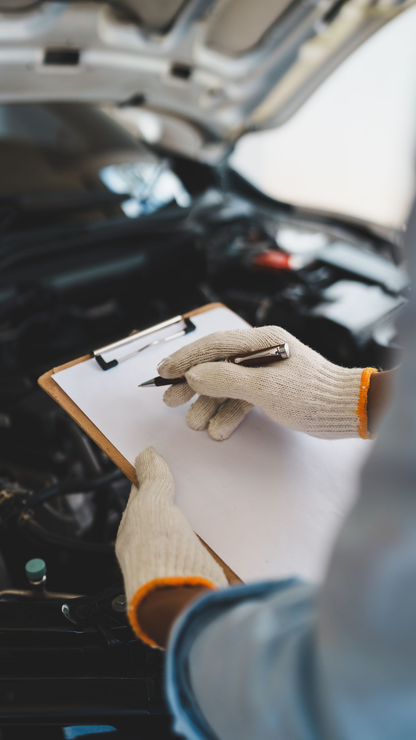 Mains gantées tenant un stylo au‑dessus d’un clipboard, moteur de voiture ouvert en arrière‑plan.