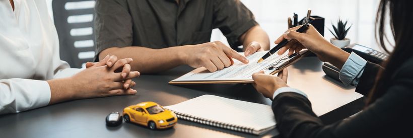 Mains pointant un document posé sur un clipboard, avec une voiture miniature sur la table.