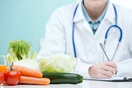 Un médico está sentado en una mesa con verduras y escribiendo en un portapapeles.