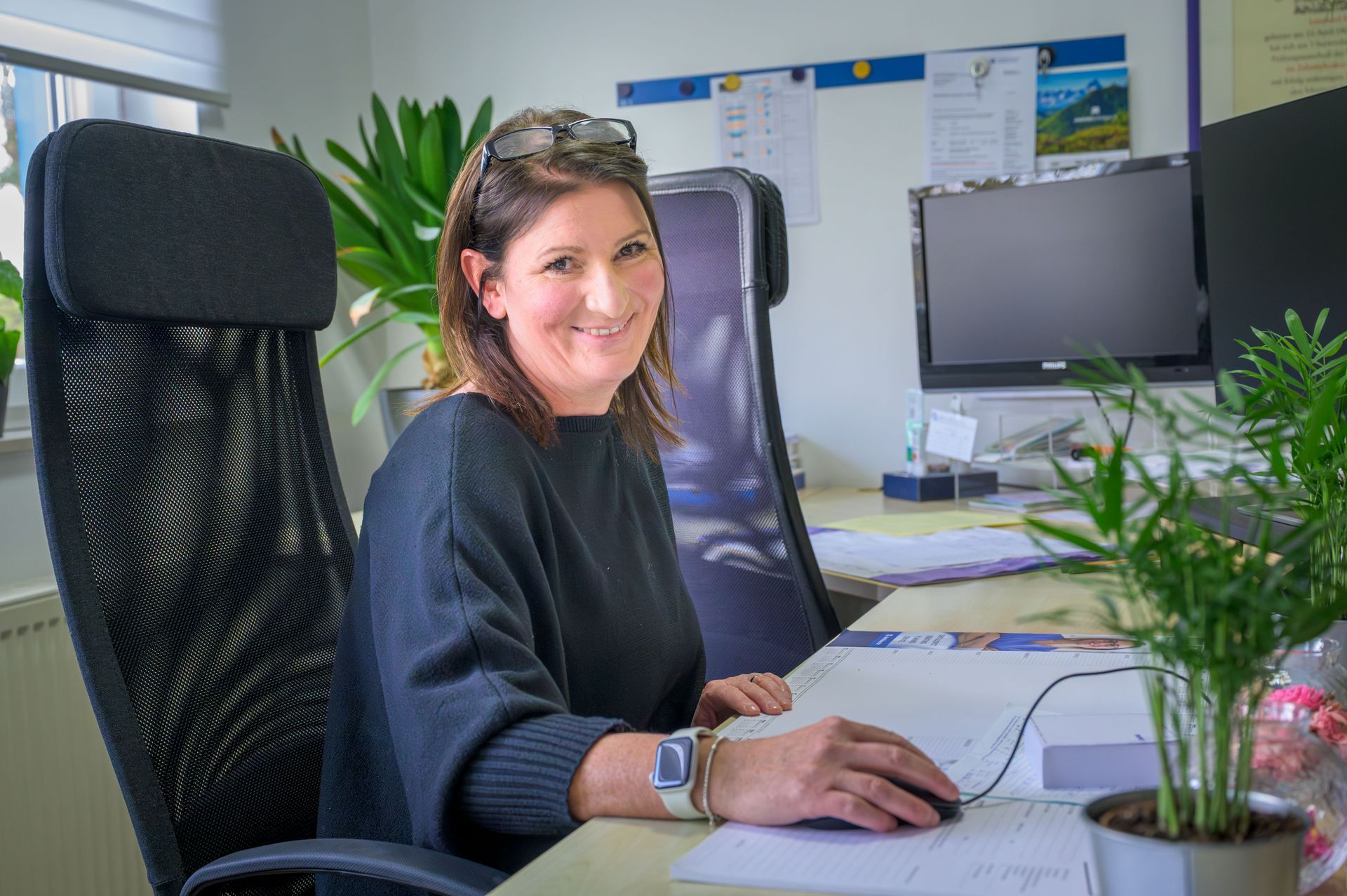Eine Frau arbeitet an einem Schreibtisch im Büro und lächelt vor einen Computer; in der Nähe befinden sich Pflanzen und Monitore.