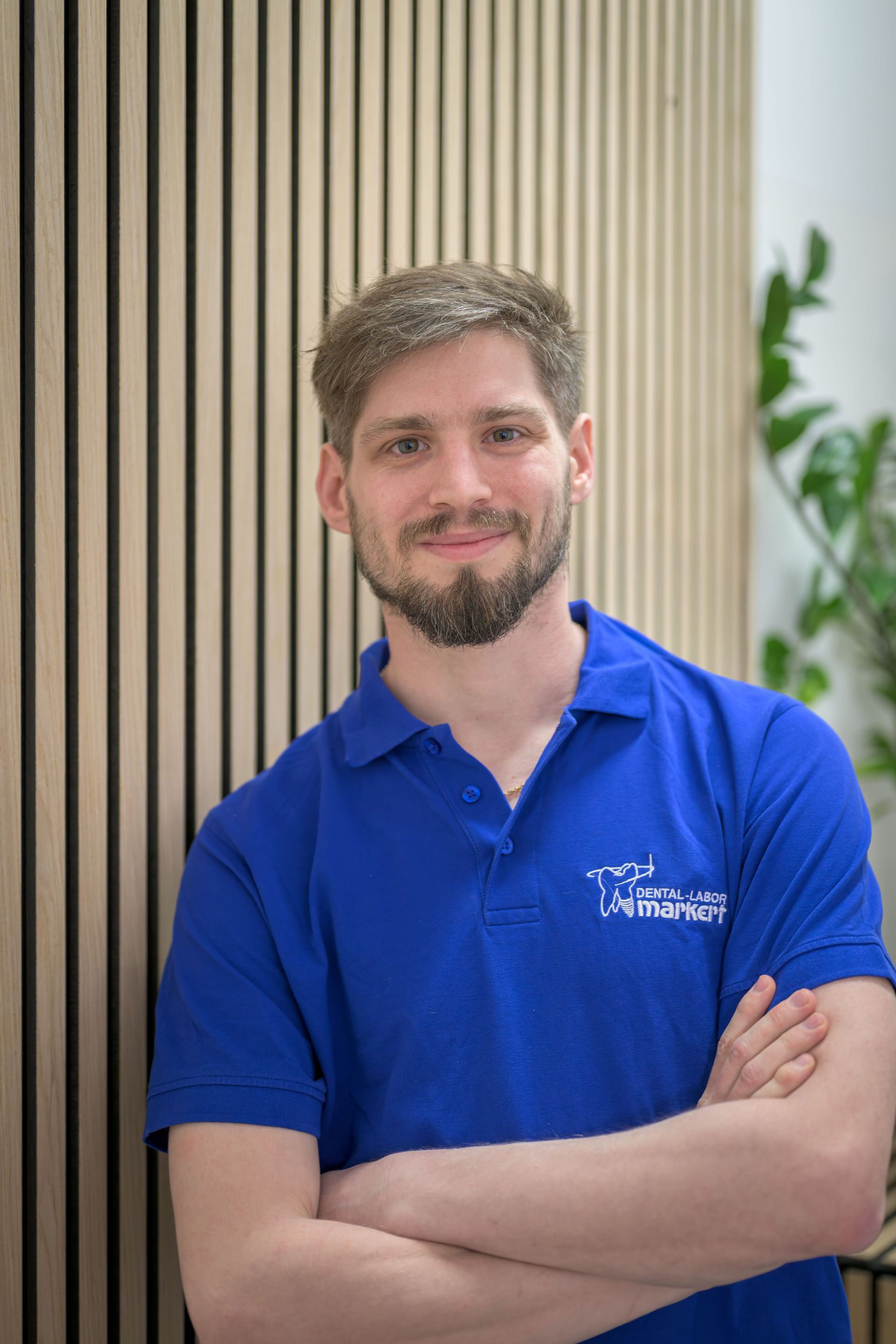 Ein lächelnder Mann in einem blauen Poloshirt mit verschränkten Armen steht vor einer gestreiften Holzwand.