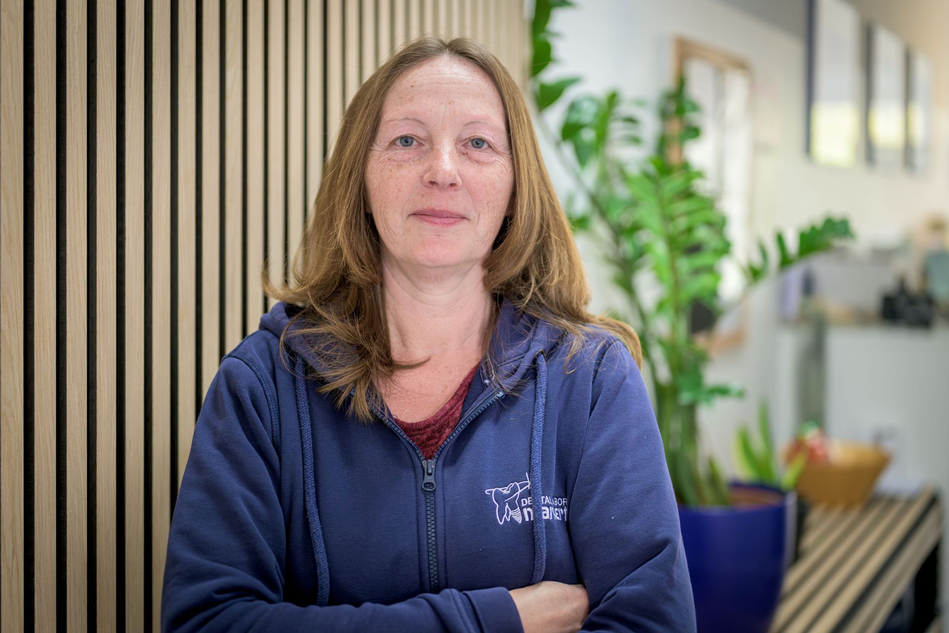 Eine Frau in einem blauen Kapuzenpullover steht mit verschränkten Armen in einem hellen Büro mit Pflanzen.