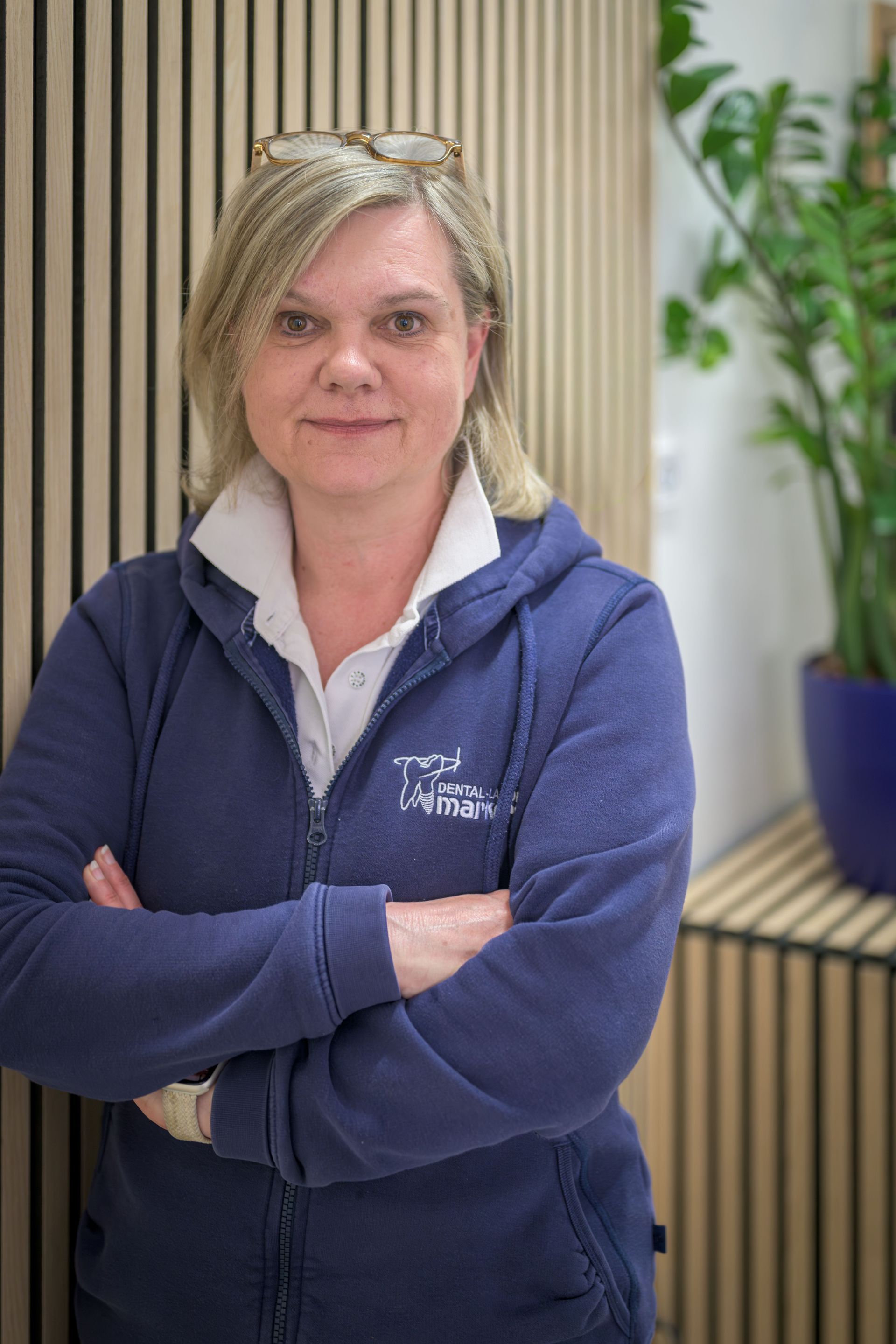 Eine Frau in einem dunkelblauen Kapuzenpulli steht mit verschränkten Armen in einem Büro, hinter ihr sind Pflanzen und Jalousien zu sehen.