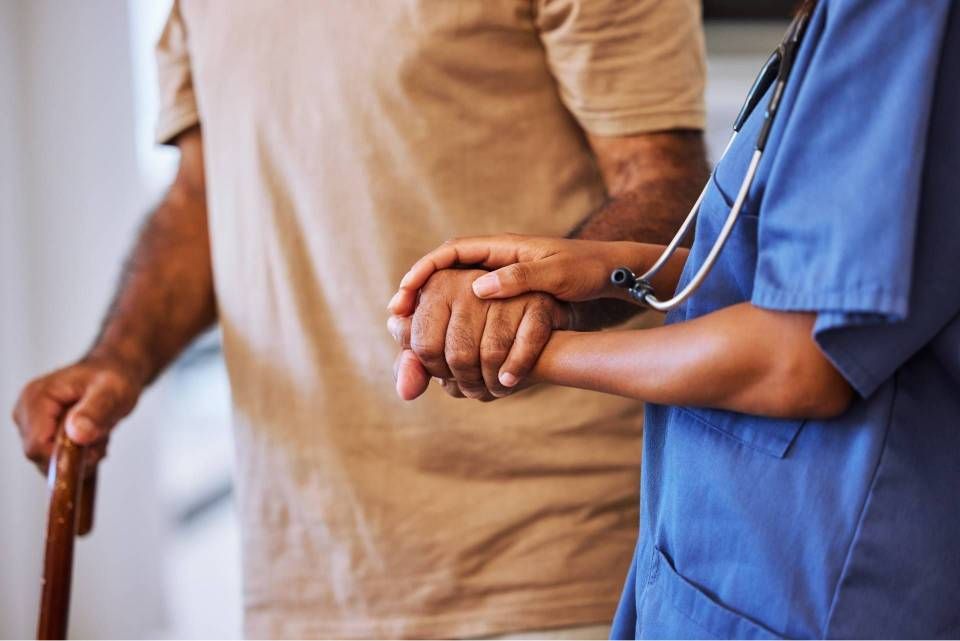 Eine Person, die einen Gehstock benutzt, wird von einer Person in blauer Uniform unterstützt, die ihre Hand hält.