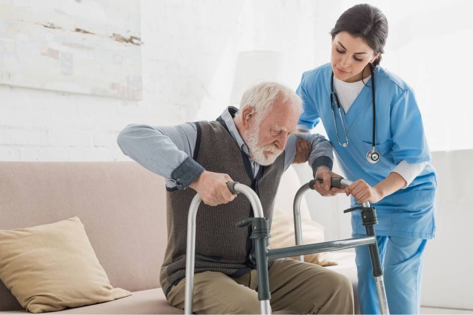 Eine Frau in blauer OP-Kleidung hilft einem älteren Mann, der einen Rollator benutzt, vom Sofa aufzustehen.