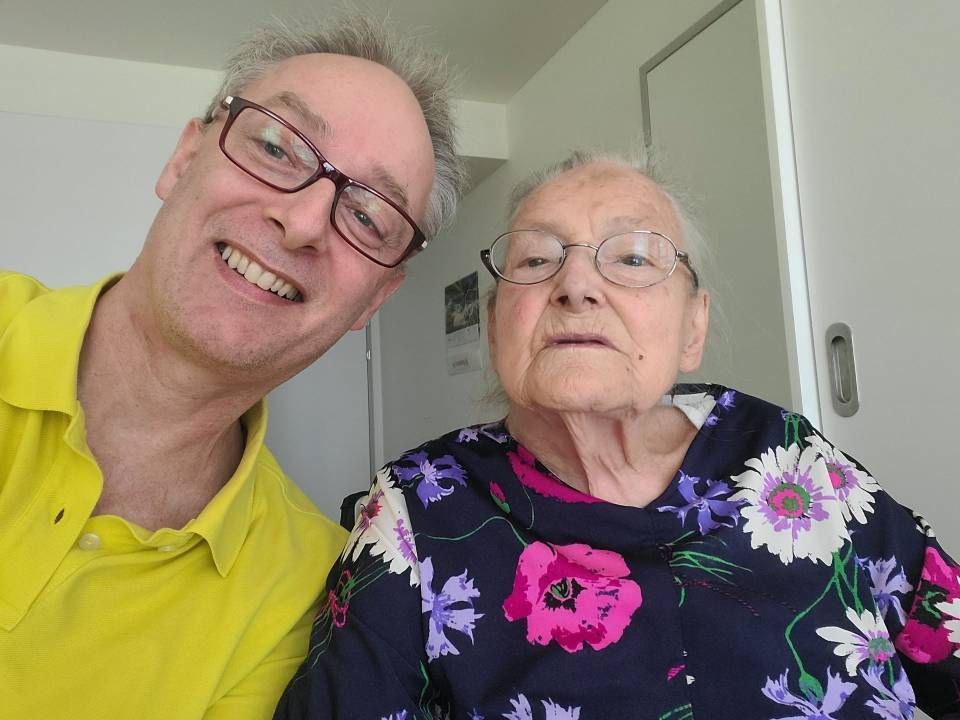 Ein Mann mit Brille lächelt und macht ein Selfie mit einer älteren Person in einem geblümten Hemd. Weiss getünchter Raum.