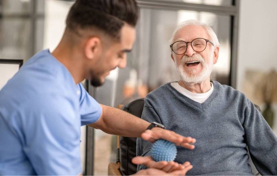 Ein Mann in OP-Kleidung hilft einer älteren Person mit einem blauen Stressball; beide lächeln.