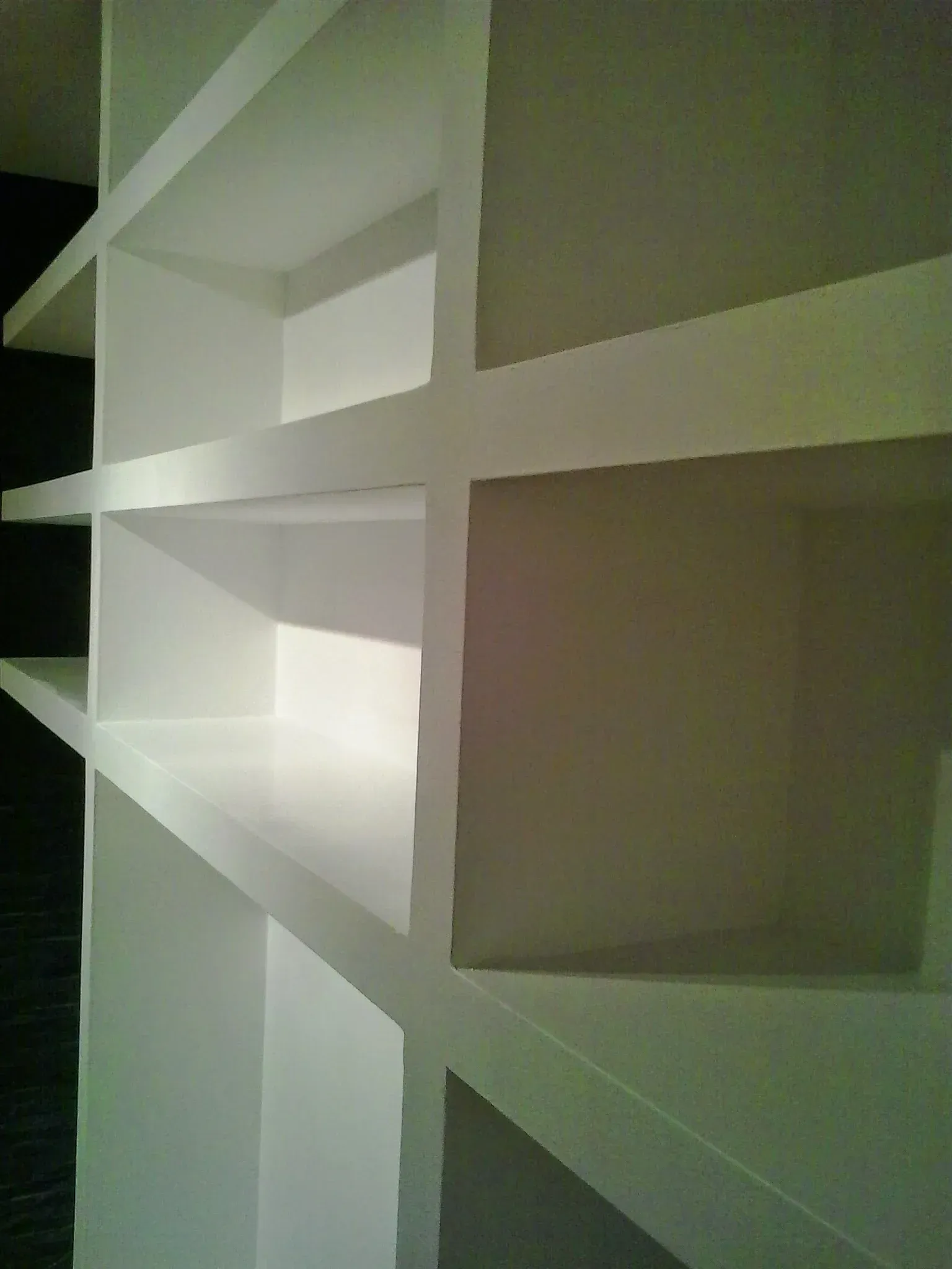 Vista lateral de estanterías empotradas blancas, vacías y con forma de compartimentos, colocadas contra una pared oscura.