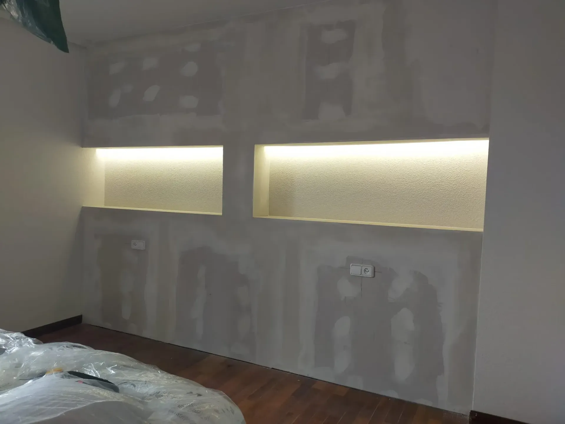 Construcción de paneles de yeso en curso, con dos nichos empotrados retroiluminados sobre un piso de madera.