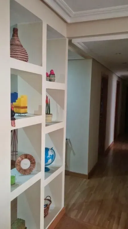 Una estantería alta y blanca, empotrada en sus compartimentos, se encuentra junto a un pasillo con suelo de madera.