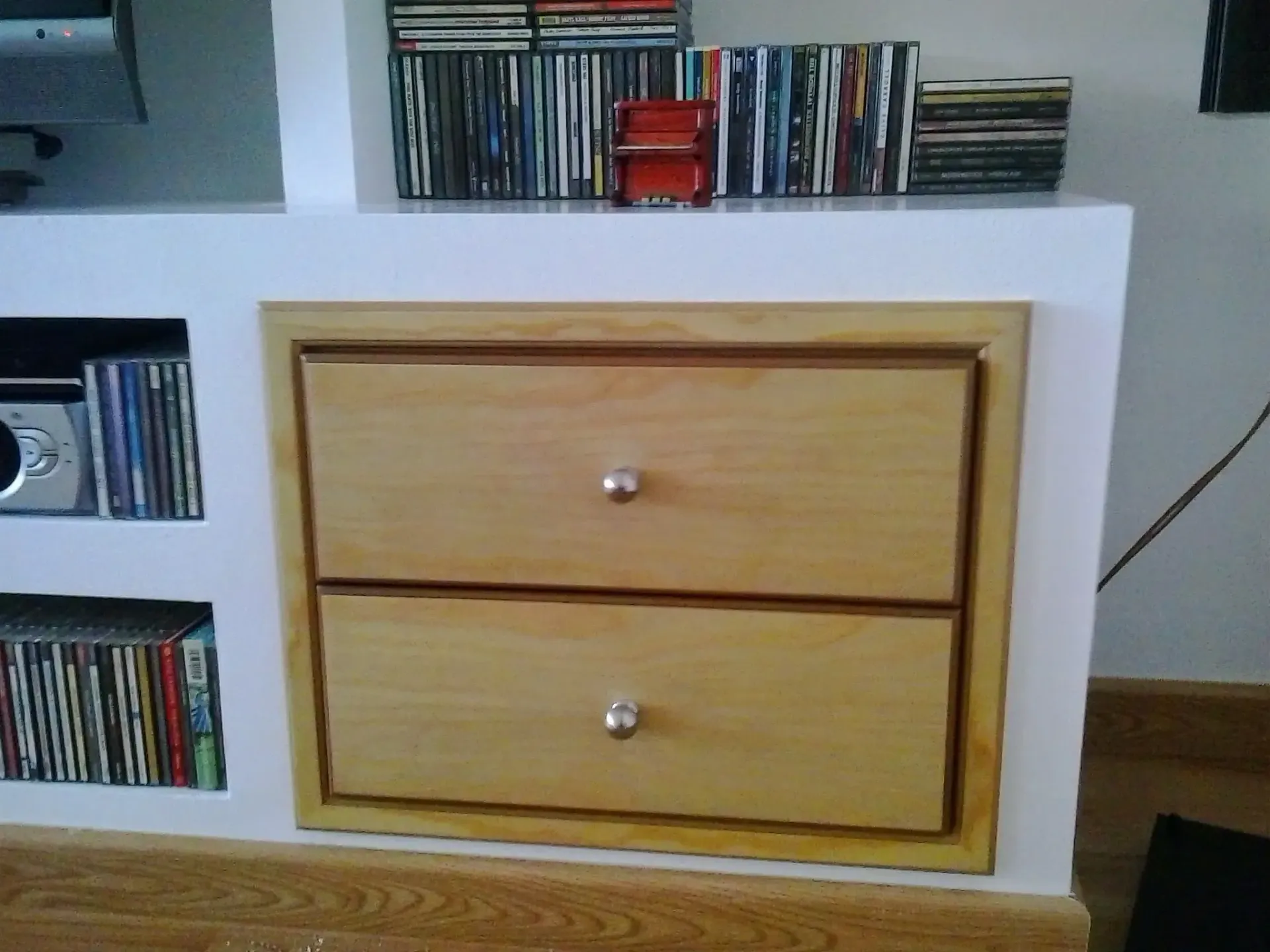 Un mueble de madera clara con dos cajones empotrado en una pared blanca, flanqueado por estantes llenos de CDs de música.