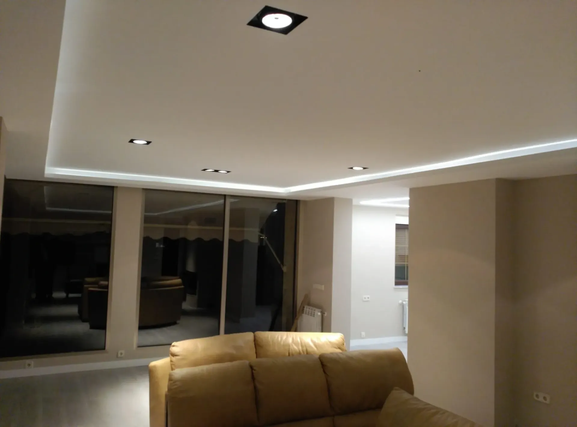 Un salón moderno por la noche, con un sofá modular de color beige, iluminación empotrada en el techo y grandes ventanales.