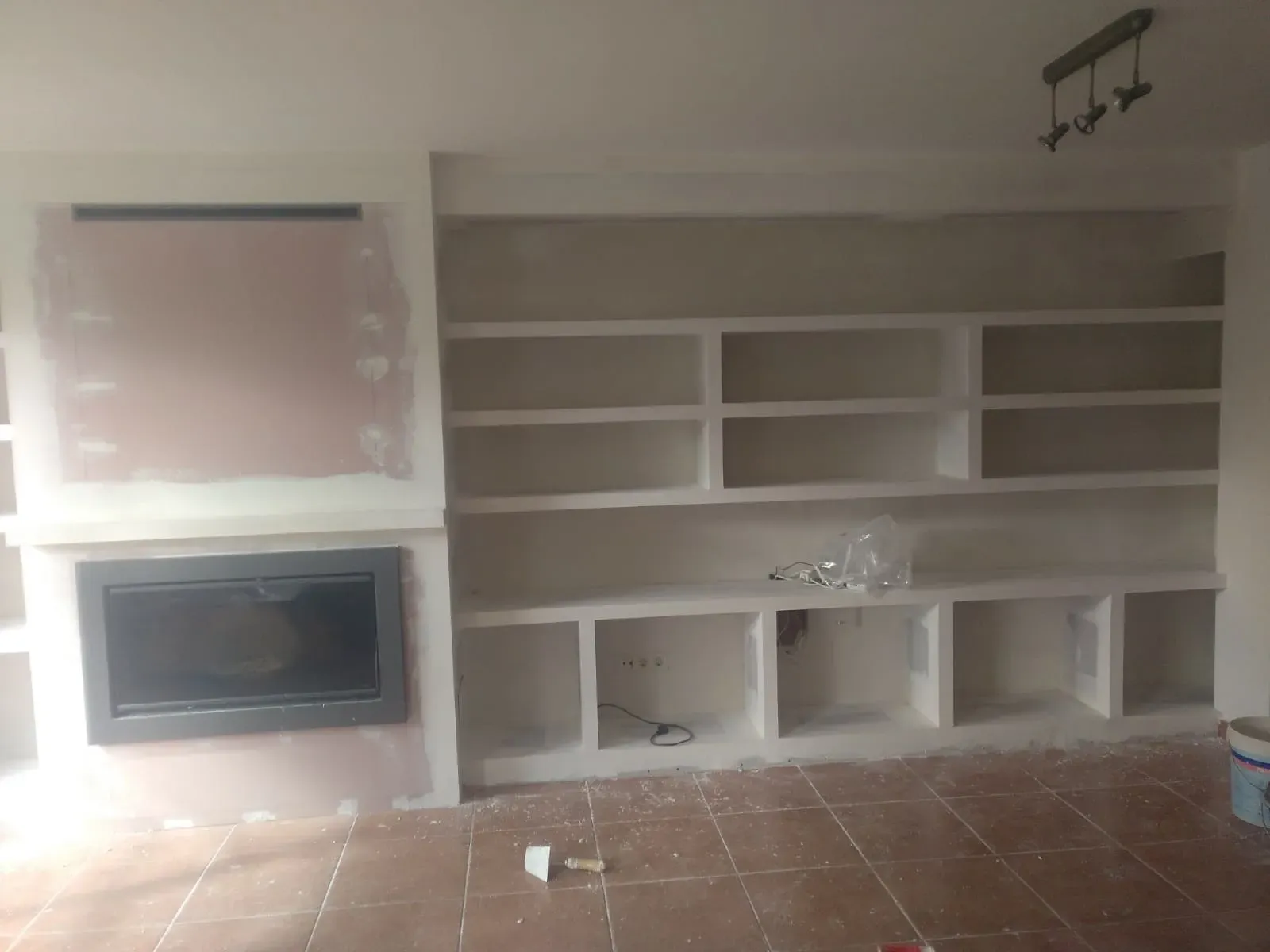 Una pared con una chimenea empotrada junto a estanterías de yeso blanco sin acabado en una habitación con suelo de baldosas.