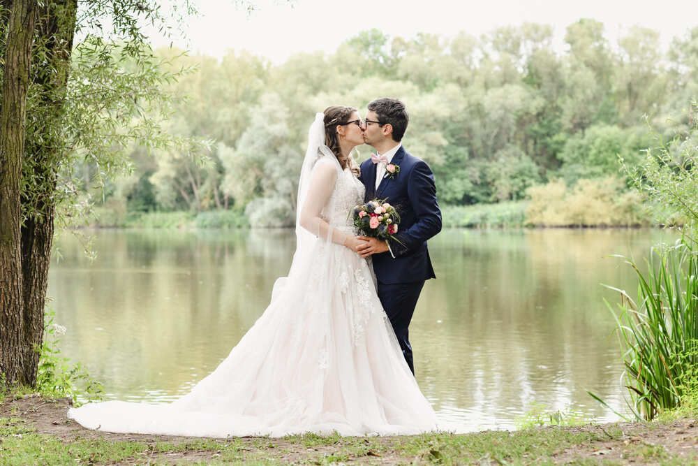 Eine Braut und ein Bräutigam küssen sich vor einem See.