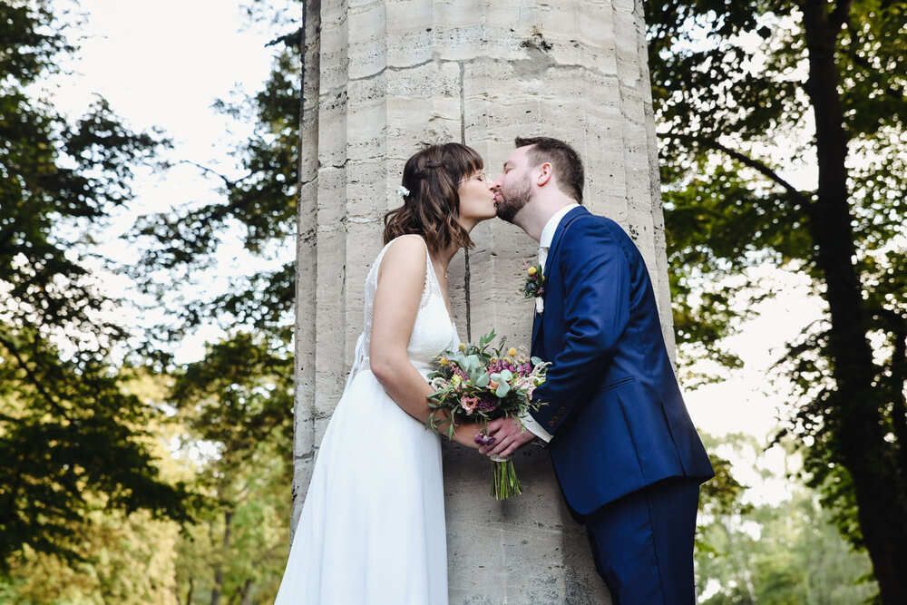 Eine Braut und ein Bräutigam küssen sich vor einer Säule.