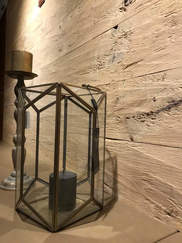 Eine Glaslaterne mit einer Kerze darin steht auf einem Tisch vor einer Holzwand.