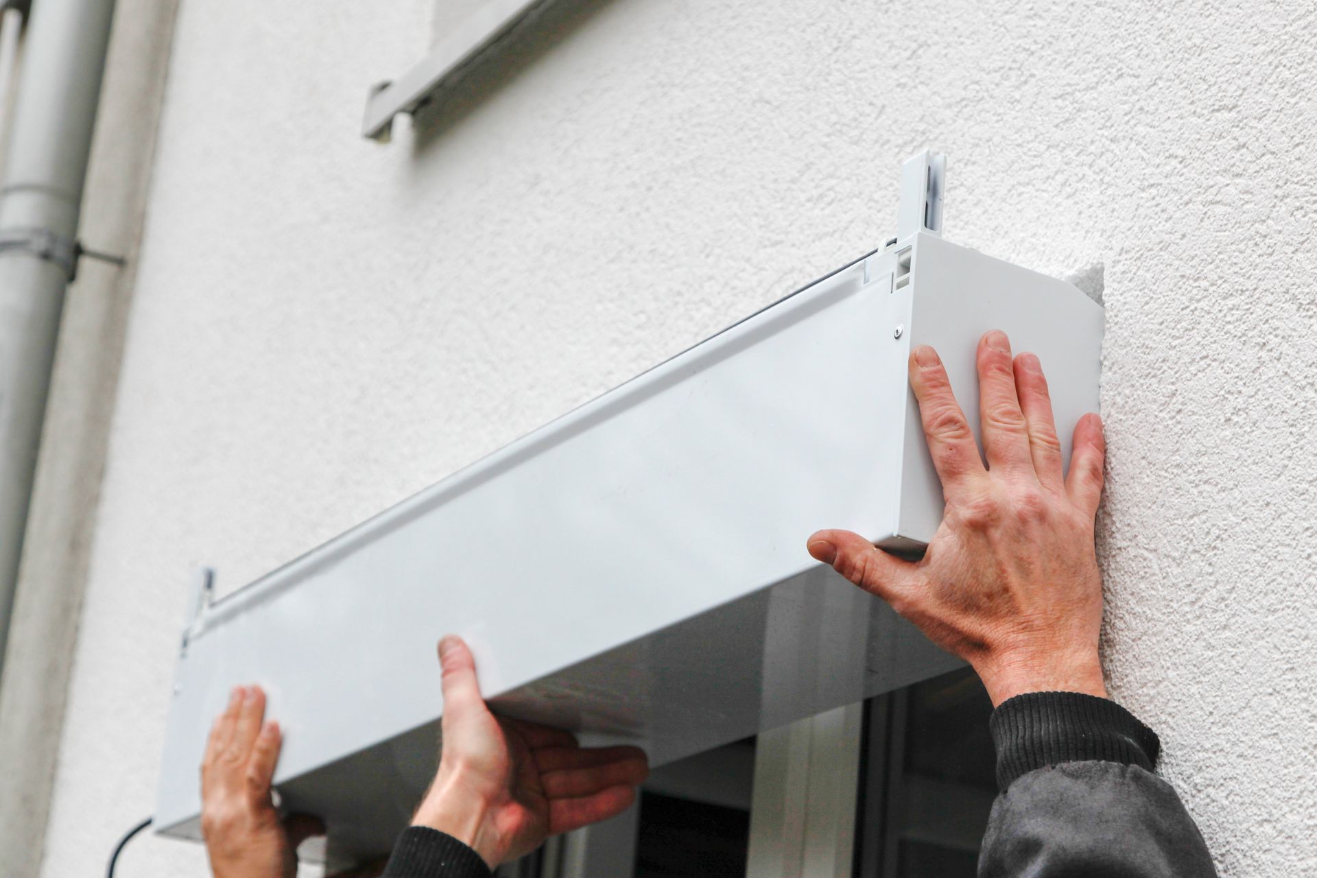 Des mains installent un auvent blanc au-dessus d’une fenêtre, sur la façade d’une maison