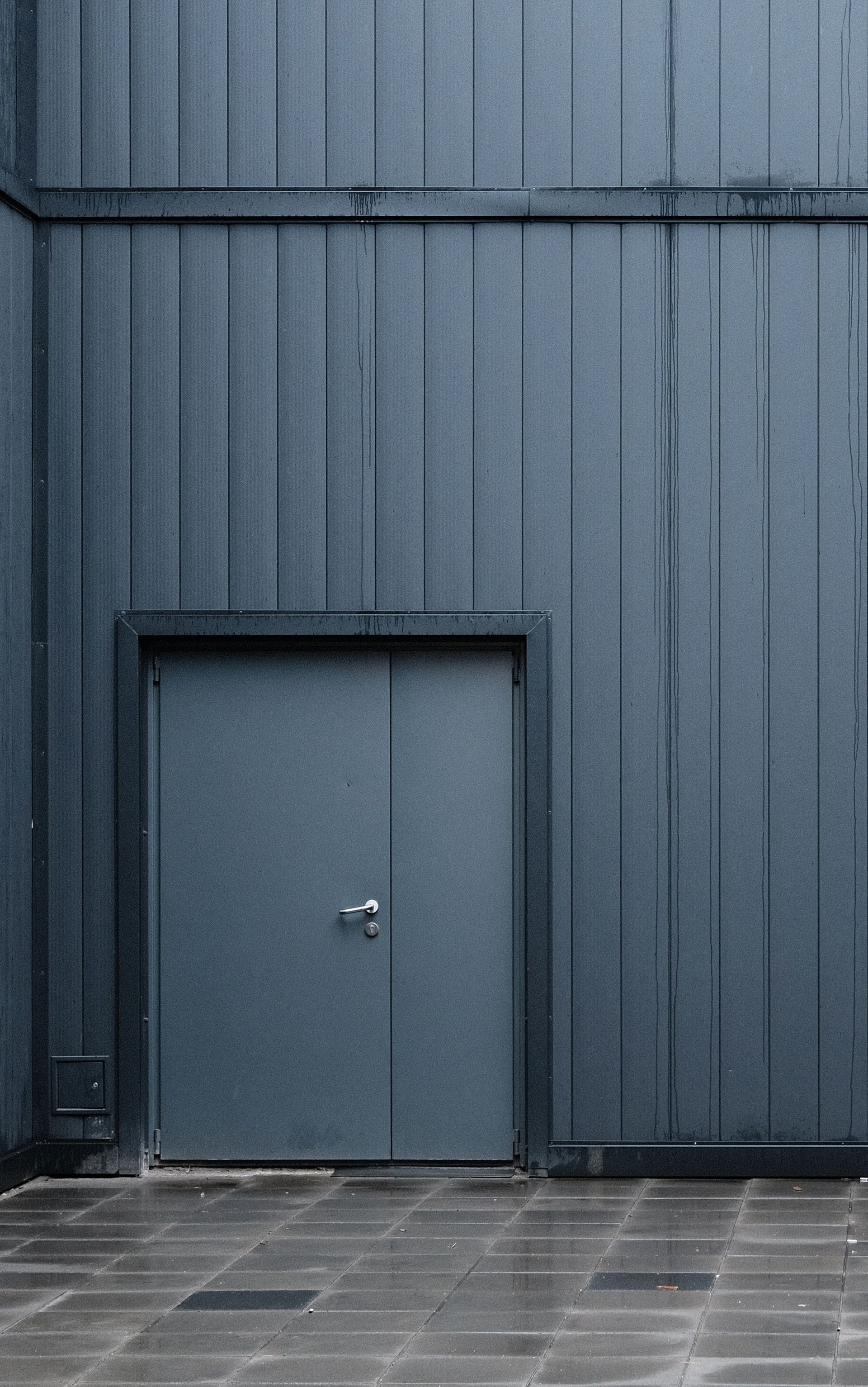 Porte métallique grise encastrée dans un mur bleu en tôle ondulée, au-dessus d’un trottoir pavé.