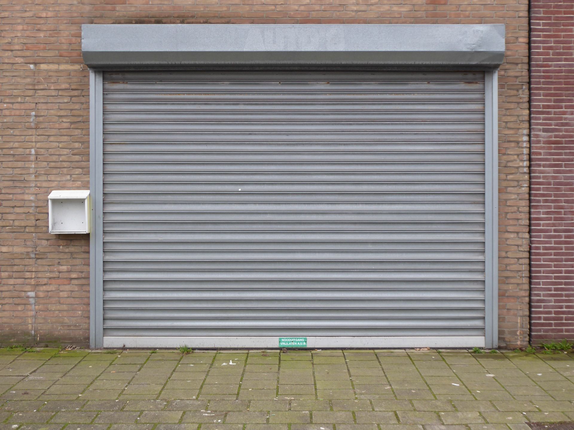 Porte de garage grise enroulable, fermée, encastrée dans un mur de briques, avec un trottoir devant