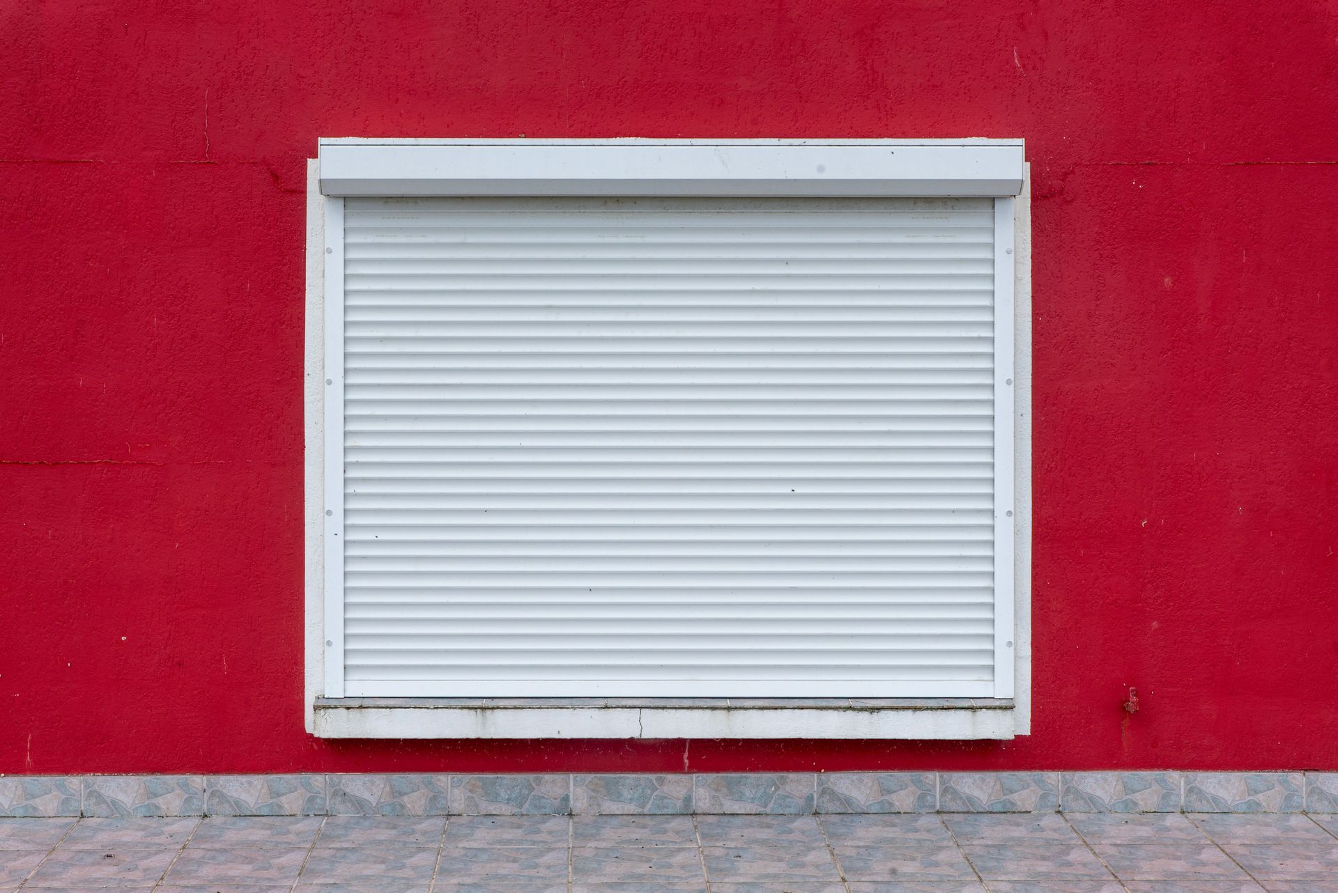 Volet blanc fermé sur un mur rouge vif avec un soubassement en pierre grise