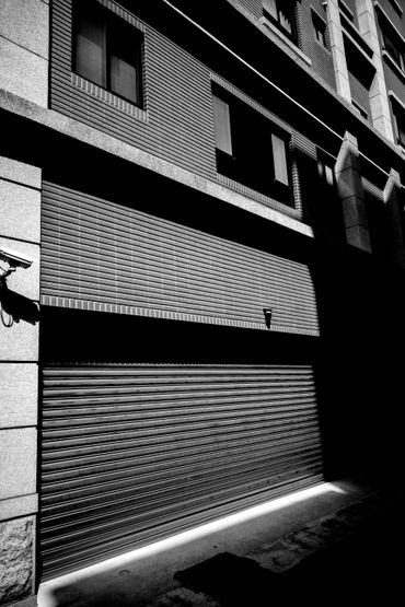 Vue en noir et blanc de la façade d’un bâtiment en briques, avec des fenêtres et un volet métallique dans une ruelle étroite