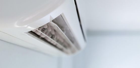 Rejilla de ventilación del aire acondicionado montada en la pared sobre un techo blanco, ligeramente abierta con polvo visible en el interior.