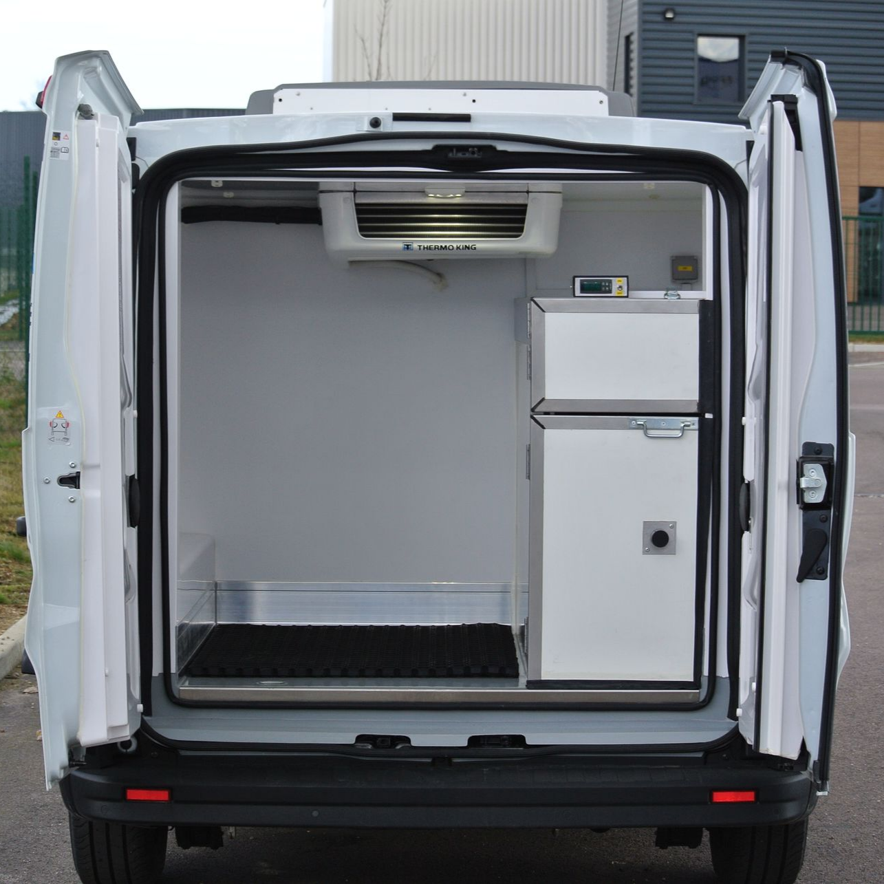 Vue arrière d'une camionnette frigorifique blanche, portes ouvertes, montrant le groupe frigorifique et les équipements intérieurs.