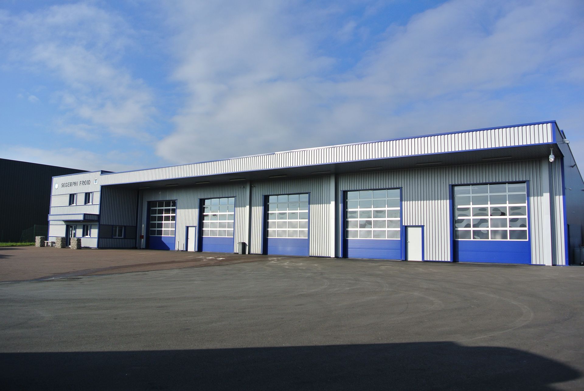 Bâtiment industriel bleu et argent avec plusieurs portes de garage sous un ciel nuageux.
