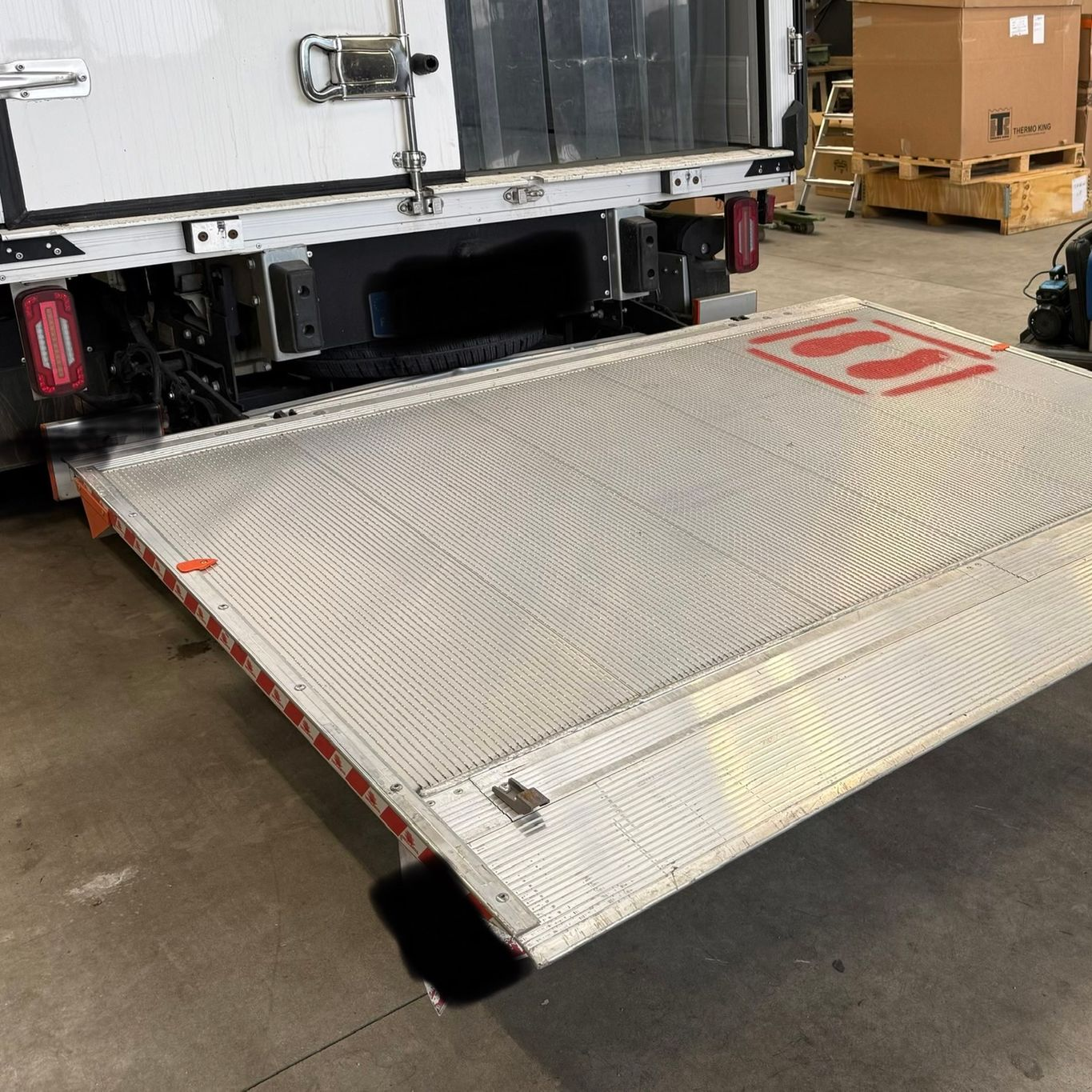 Vue arrière d'un camion de livraison blanc avec un hayon élévateur argenté déployé jusqu'au sol, à l'intérieur d'un entrepôt.