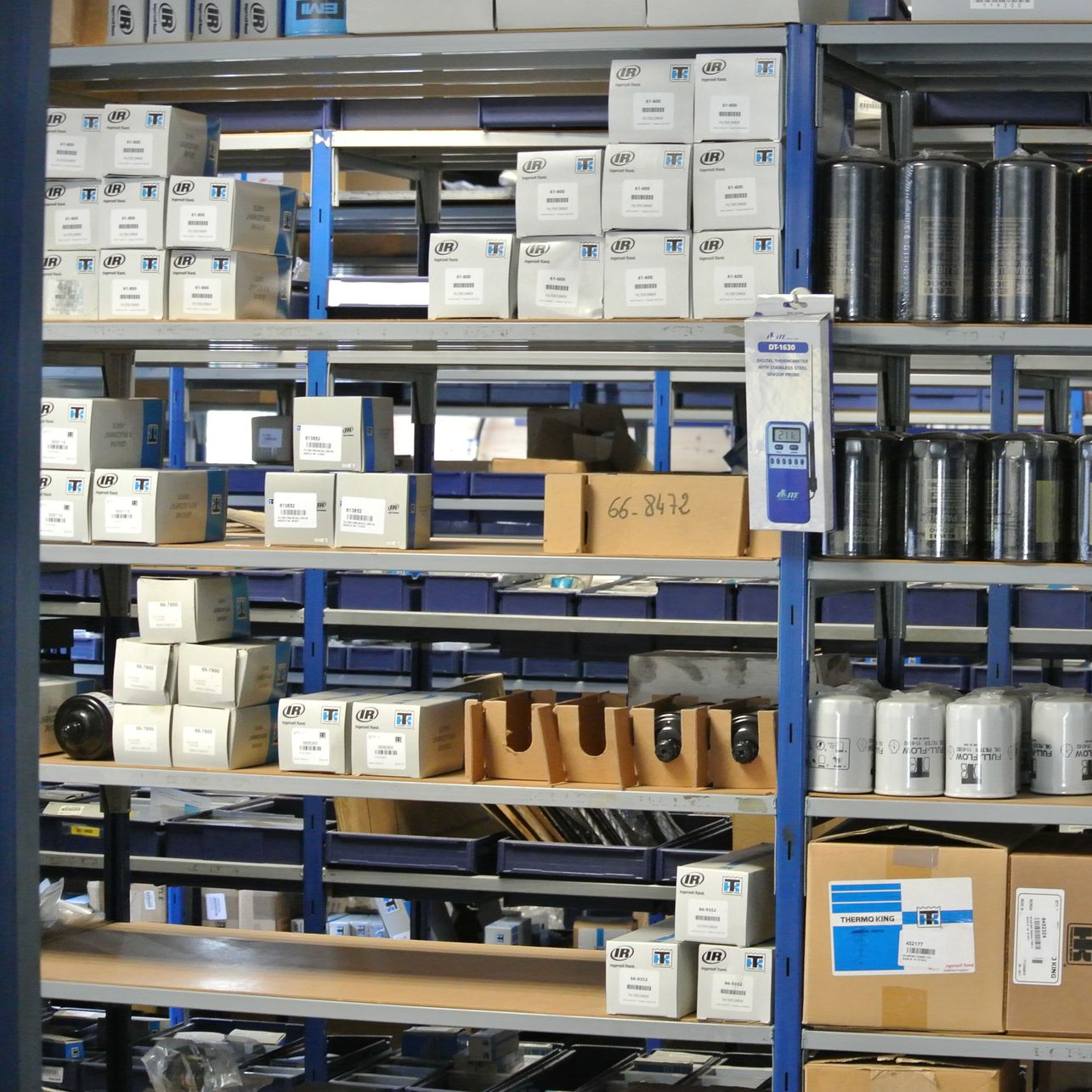 Des étagères remplies de boîtes et de pièces détachées dans un entrepôt, quelques bacs bleus.