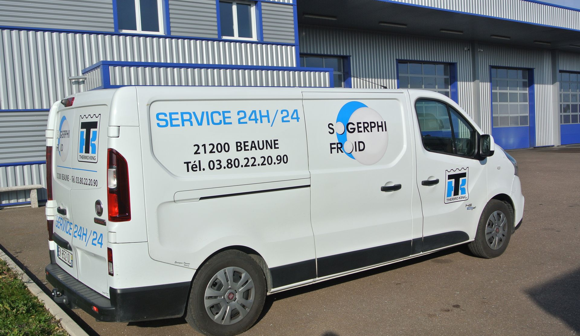 Fourgonnette de service blanche avec l'inscription SERVICE 24H/24, stationnée devant un bâtiment.