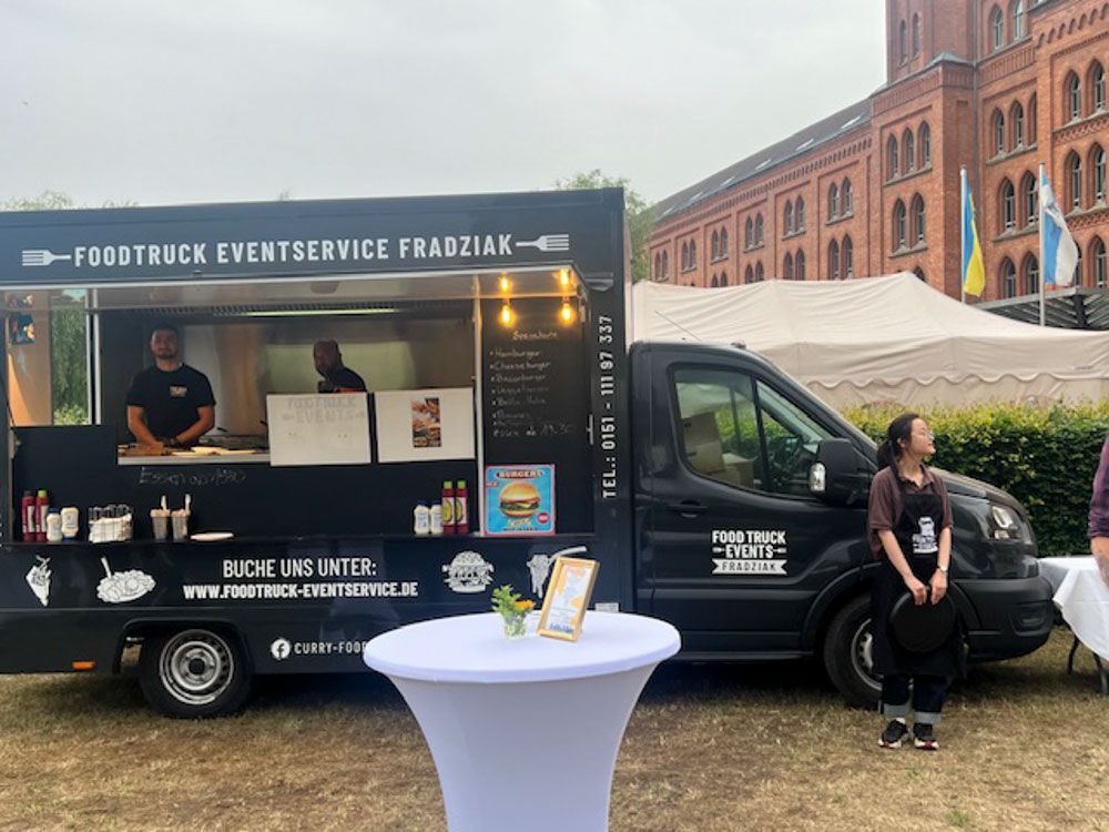 Vor einem Gebäude parkt ein Imbisswagen, davor steht ein Tisch.