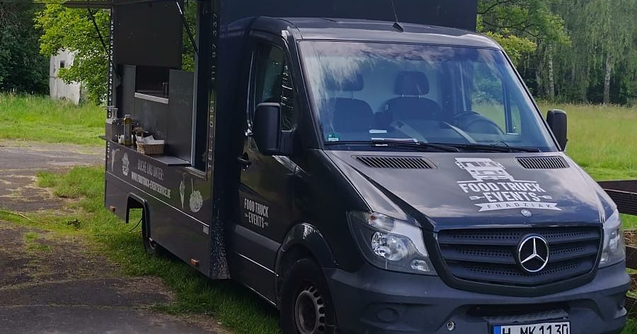 Schwarzer Mercedes-Imbisswagen parkt auf einer Rasenfläche; das Ausgabefenster ist geöffnet und das Logo befindet sich an der Vorderseite.