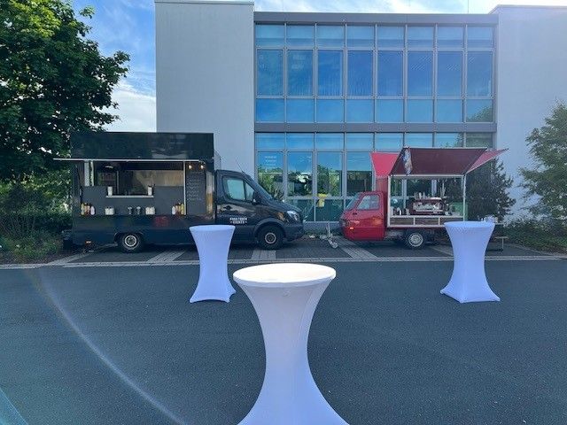 Vor einem Gebäude parken zwei Imbisswagen mit Tischen davor.